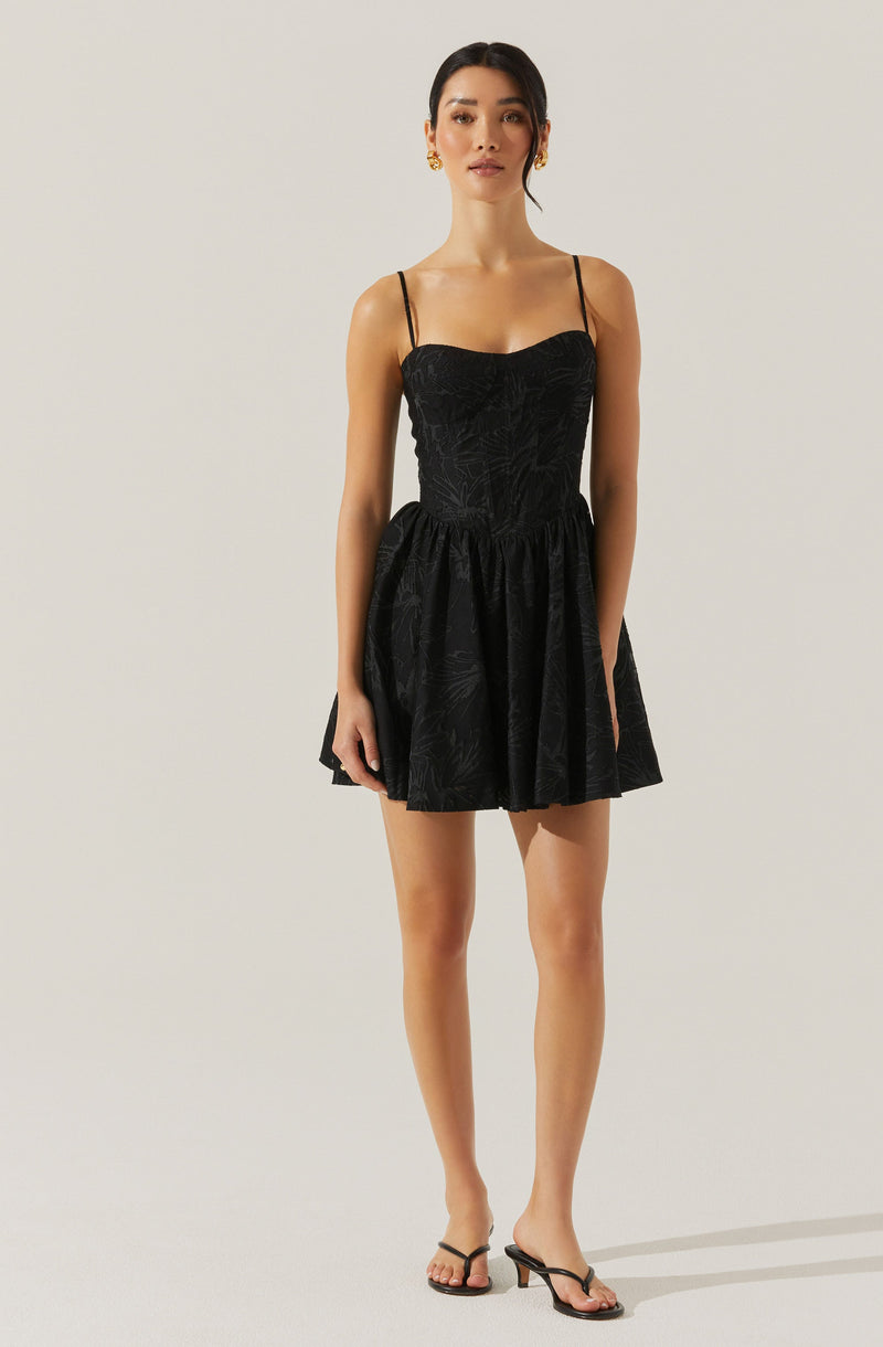A woman is wearing a black dress with a fitted bodice and thin spaghetti straps. The dress has a subtle textured pattern and a full, gathered skirt that falls above the knees. She is accessorizing with gold earrings and black heeled sandals.