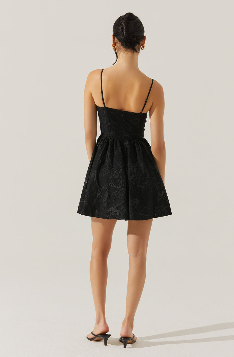 The woman is wearing a black dress with thin spaghetti straps. The dress has a fitted bodice and a full, slightly flared skirt with subtle floral patterns. She is also wearing small earrings and black heeled sandals.