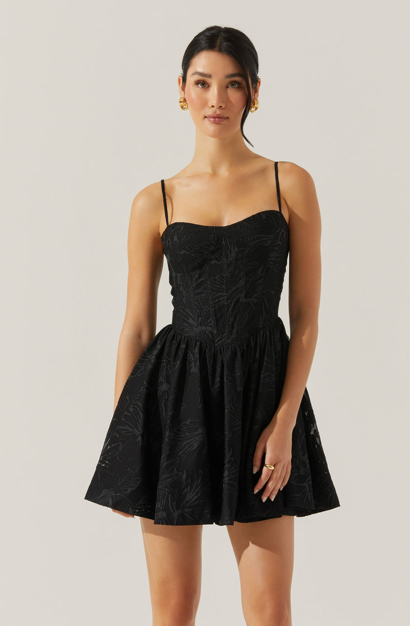 A woman is wearing a black sleeveless dress with thin spaghetti straps. The dress features a fitted bodice with subtle textured patterns and a flared, knee-length skirt. The fabric has a faint, leafy or botanical print. She is accessorized with gold earrings and a ring.