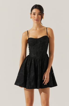 A woman is wearing a black sleeveless dress with thin spaghetti straps. The dress features a fitted bodice with subtle textured patterns and a flared, knee-length skirt. The fabric has a faint, leafy or botanical print. She is accessorized with gold earrings and a ring.