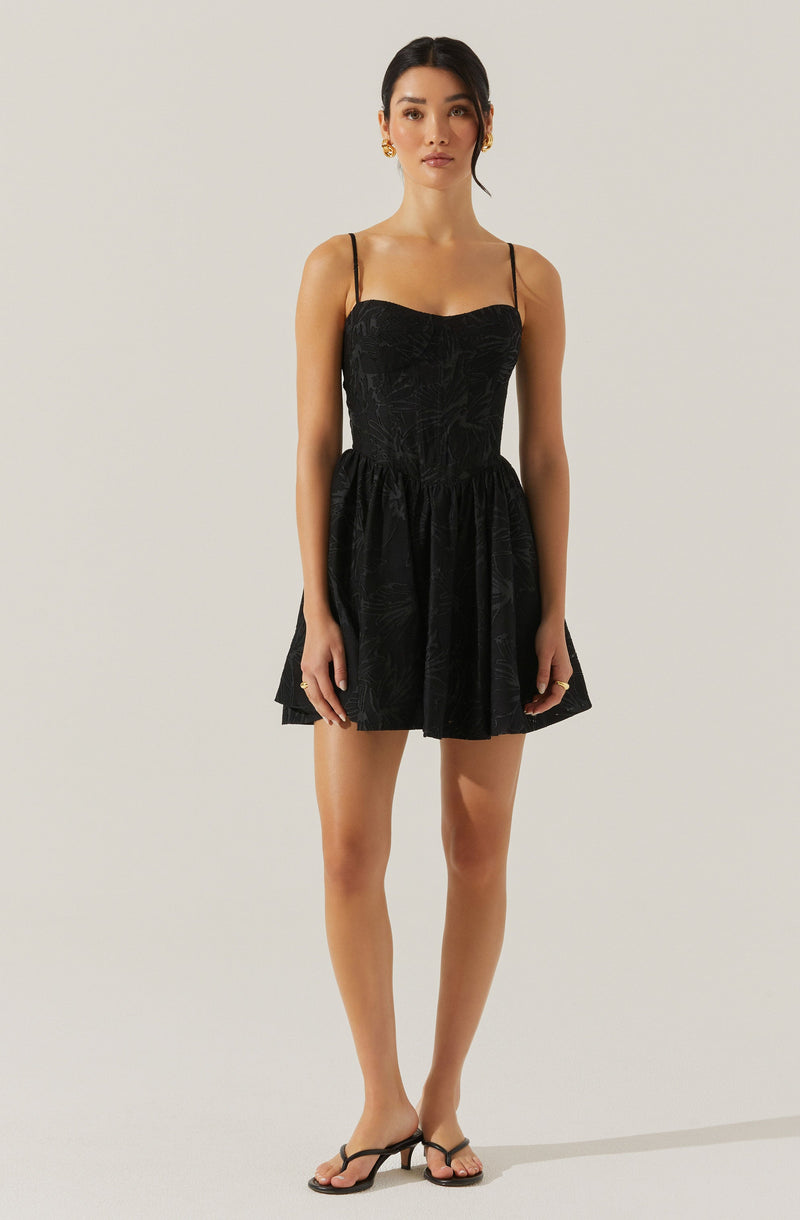 A woman is wearing a black dress with thin spaghetti straps. The dress has a fitted bodice with a slightly ruched design and a flared, above-the-knee skirt. She is also wearing black open-toe sandals with a small heel. Her accessories include gold earrings and rings. Her hair is styled in a sleek, low bun, and she has a neutral facial expression.