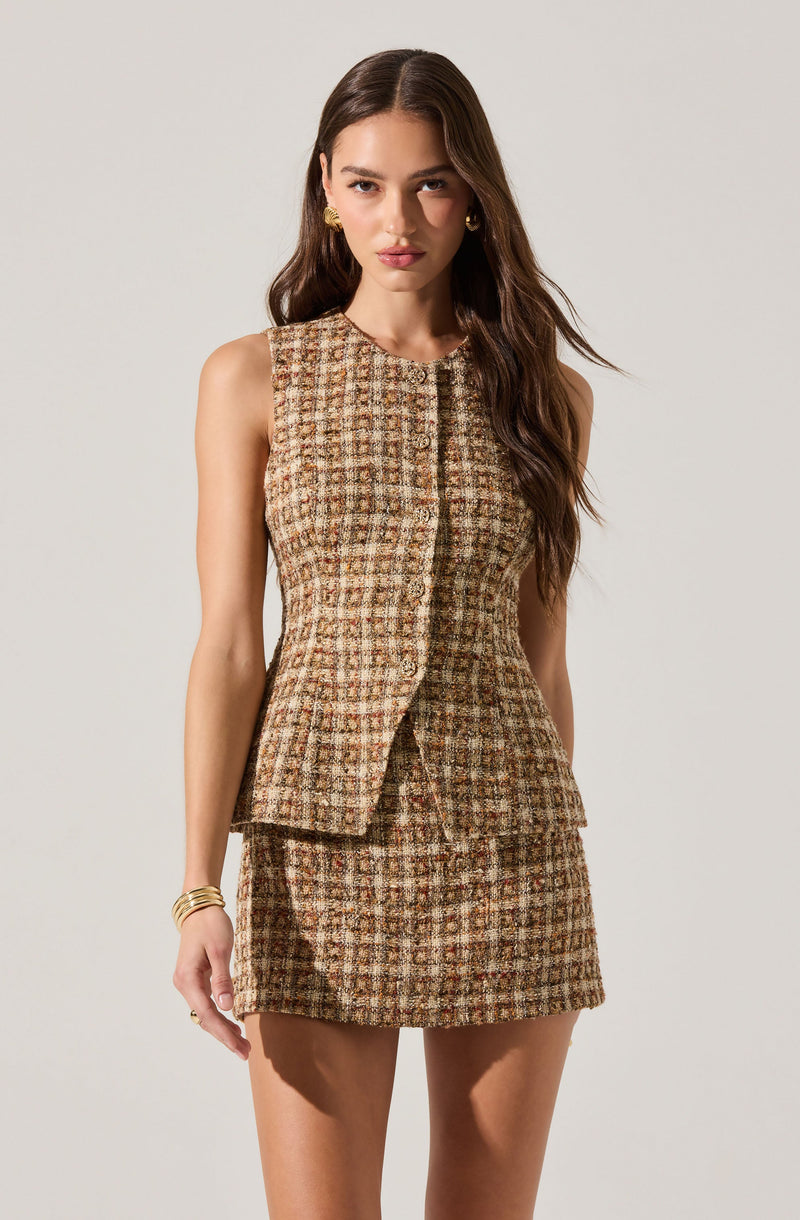 A woman is wearing a sleeveless, fitted top and skirt set made of a textured, plaid fabric in shades of brown, beige, and green. The top has a structured, tailored fit with a round neckline and a front opening that appears to be fastened with buttons. The skirt is short and fitted, matching the fabric of the top. The overall outfit has a classic, sophisticated look with a checkered pattern.