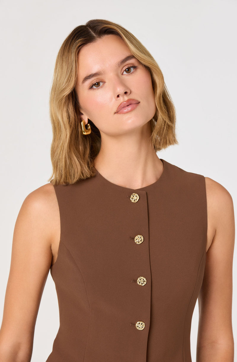 A woman with shoulder-length wavy blonde hair is wearing a sleeveless brown dress with a round neckline. The dress features a row of decorative gold buttons running down the front. She has on large, gold hoop earrings.