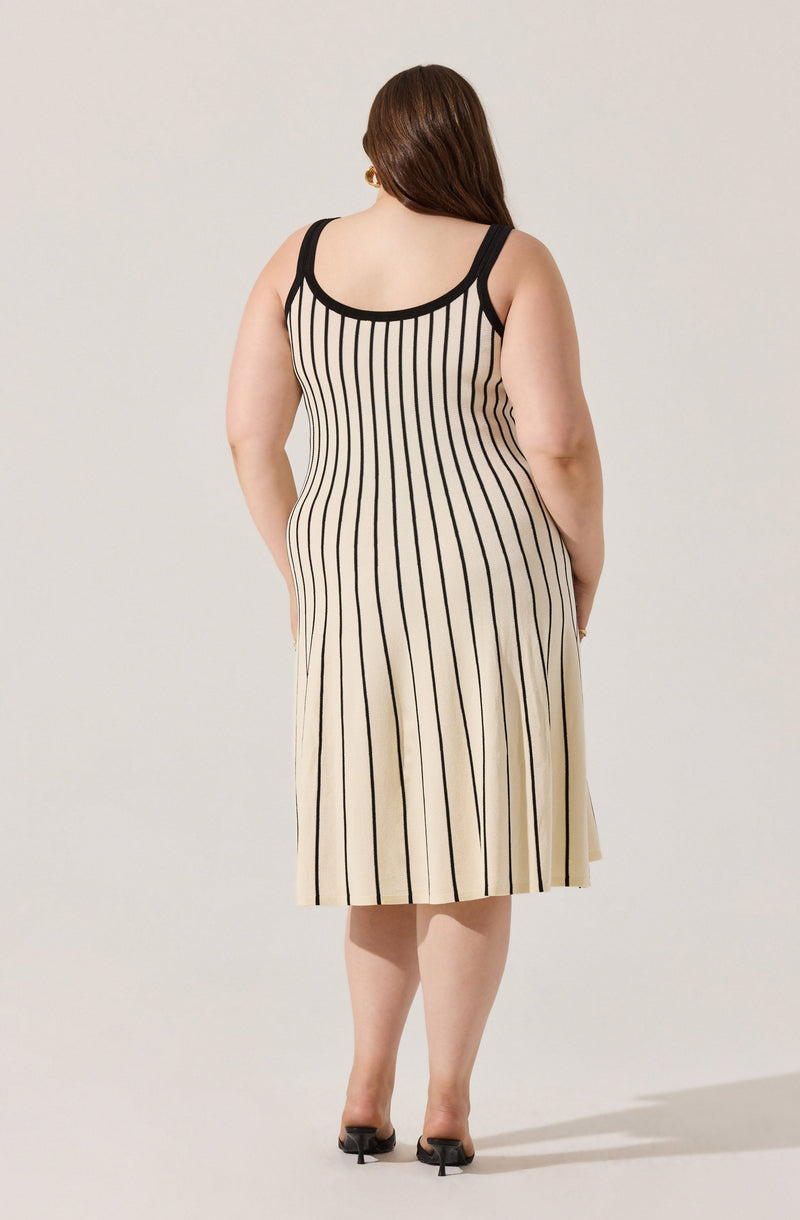 A woman is wearing a sleeveless, knee-length dress with black vertical stripes on a beige background. The dress has black trim around the neckline and armholes. She is also wearing black open-toed heels.
