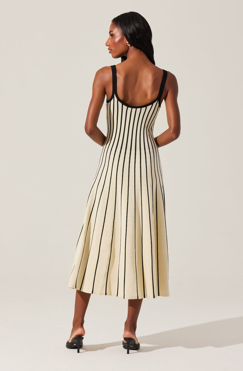 A woman is wearing a sleeveless, cream-colored dress with black vertical stripes. The dress has black shoulder straps and a fitted bodice that flares out into a long, flowing skirt reaching mid-calf. She is paired with black open-toe heels.