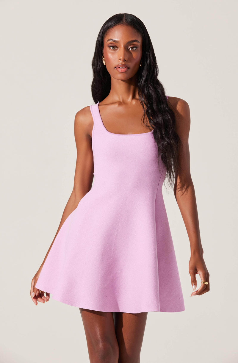 A woman is wearing a sleeveless, light purple dress with a fitted bodice and a flared skirt that falls above the knees. The dress has wide shoulder straps and a square neckline.