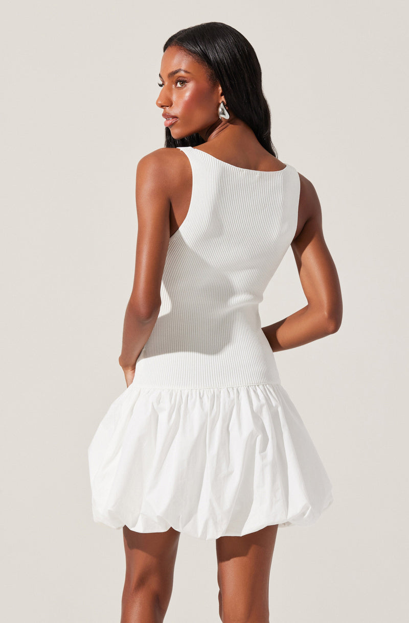 A woman is wearing a sleeveless, white ribbed knit top with a wide, rounded neckline. She is also wearing a white, voluminous skirt with gathered fabric that creates a bubble or puffy appearance at the hem. She has long, straight black hair and is accessorized with large, statement earrings.