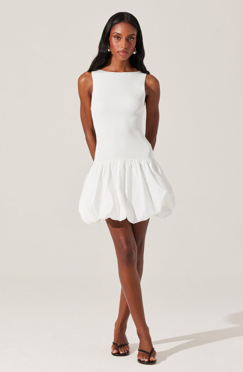 A woman is wearing a sleeveless, knee-length white dress with a ribbed bodice and a wide, gathered, bubble-style skirt. She accessorizes with earrings and black high-heeled sandals.