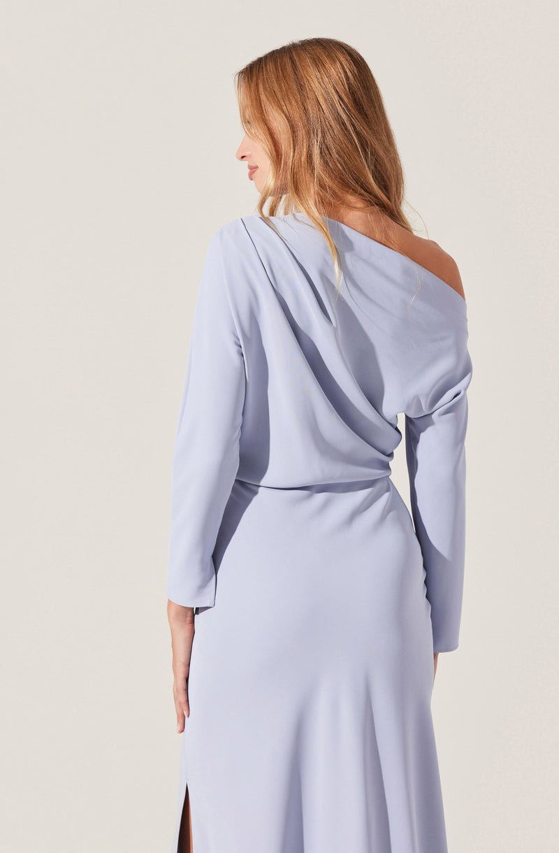 A woman with light brown hair is wearing a light blue, long-sleeved dress. The dress features a draped design on the upper part, with an off-the-shoulder style on one side, and a fitted waist. The fabric appears smooth and slightly shiny, flowing down to a long skirt with a small slit on one side.