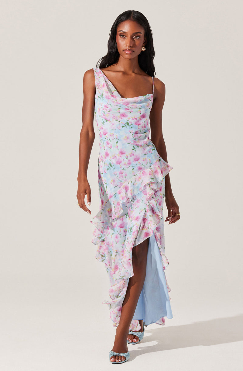 A woman is wearing a sleeveless, asymmetrical dress with spaghetti straps. The dress features a pastel watercolor floral pattern in shades of pink, blue, and green. It has ruffled details along the hemline and front slit, revealing a light blue underlayer. She is also wearing open-toe heels with a crisscross design.