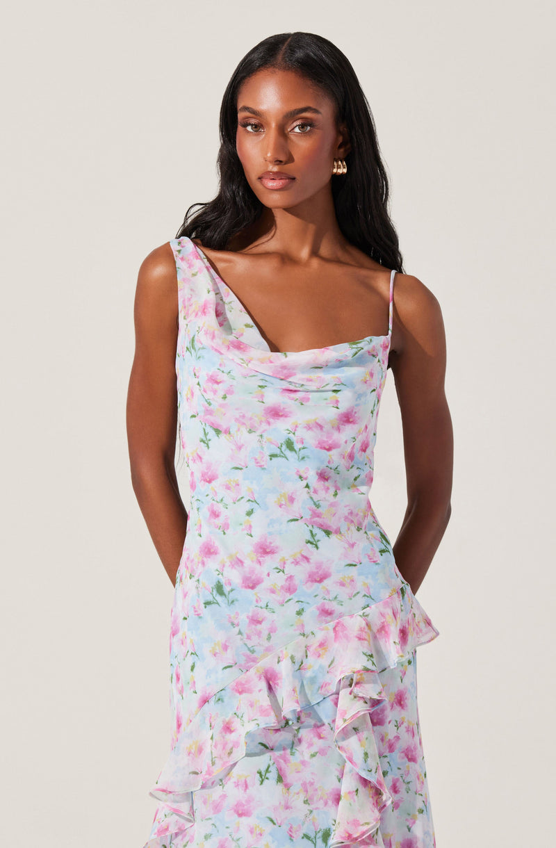 The dress features a pastel floral pattern with pink, purple, and green colors. It has thin spaghetti straps and a draped neckline that creates a soft, elegant look. The fabric flows smoothly and includes ruffled details along the hemline, adding a touch of femininity and movement to the overall design.