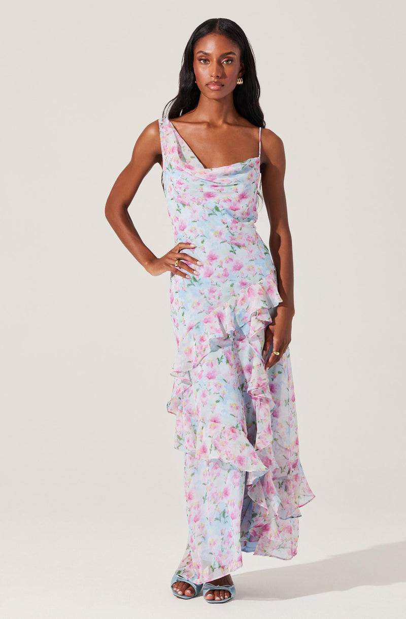 A woman is wearing a sleeveless, ankle-length dress with a pastel floral pattern. The dress features a cowl neckline, thin spaghetti straps, and ruffled details along the front and hem. She is also wearing open-toe high-heeled sandals.