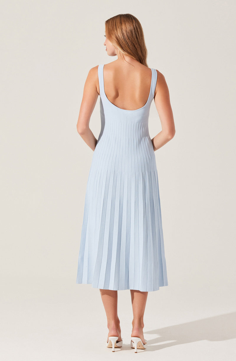 A woman is wearing a light blue, sleeveless, midi-length dress with wide shoulder straps. The dress features vertical pleats throughout the fabric. She is also wearing white high-heeled shoes.