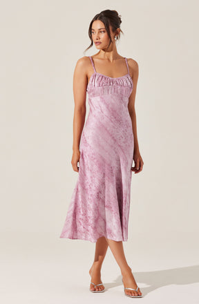 A woman is wearing a pink, velvety midi dress with a fitted bodice and thin spaghetti straps. The dress features a gathered neckline with a ruffle detail across the chest. The fabric has a subtle textured pattern and a soft sheen, creating an elegant and luxurious appearance. She is paired with transparent high heels.