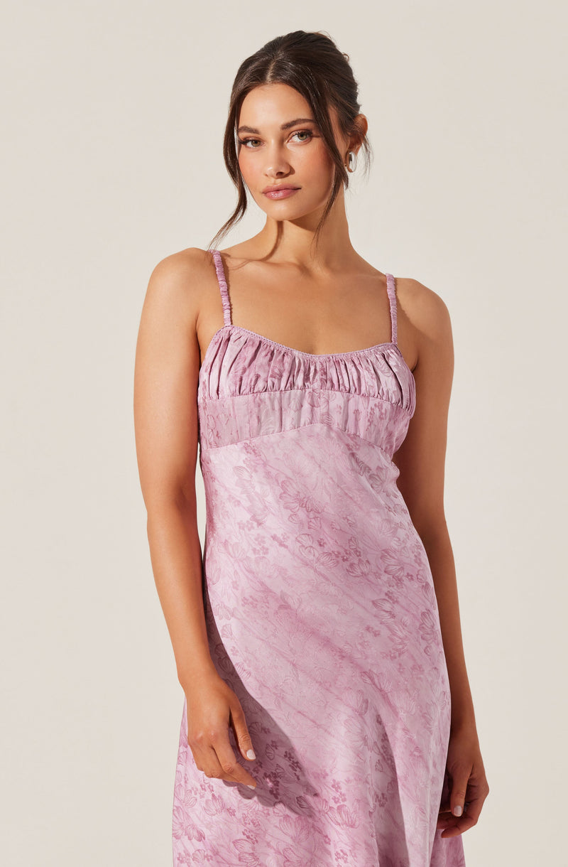 A woman is wearing a sleeveless, pink dress with thin spaghetti straps. The dress features a gathered, ruched bodice with a slightly textured, floral-inspired pattern throughout the fabric.