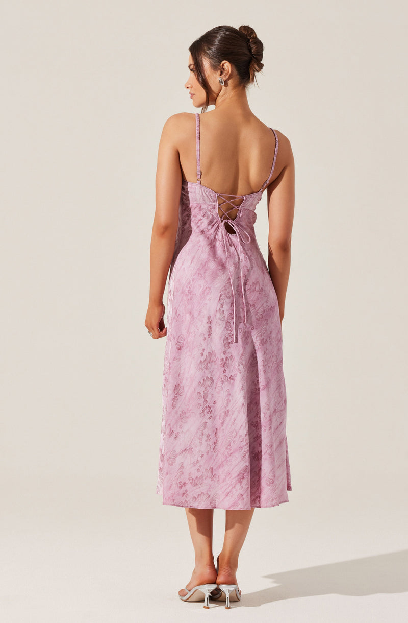 A woman is wearing a pink dress with a subtle floral pattern. The dress has thin straps, a low-cut back with crisscross lacing, and ties at the waist. The length of the dress reaches mid-calf. She is also wearing light-colored heeled shoes.