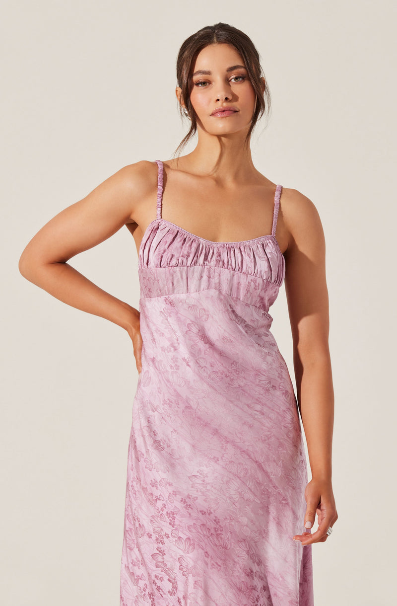 A woman is wearing a light purple satin slip dress with spaghetti straps. The dress features a gathered bodice with ruching details and a subtle floral pattern throughout the fabric.