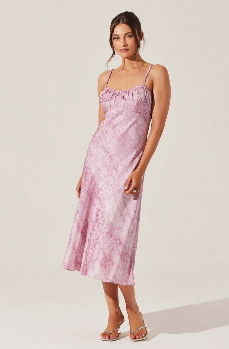 A woman is wearing a sleeveless, ankle-length dress with thin spaghetti straps. The dress has a pink, shiny, textured fabric with a subtle pattern, and features a gathered, ruffled detail at the top. She is also wearing open-toe, high-heeled sandals with clear straps.