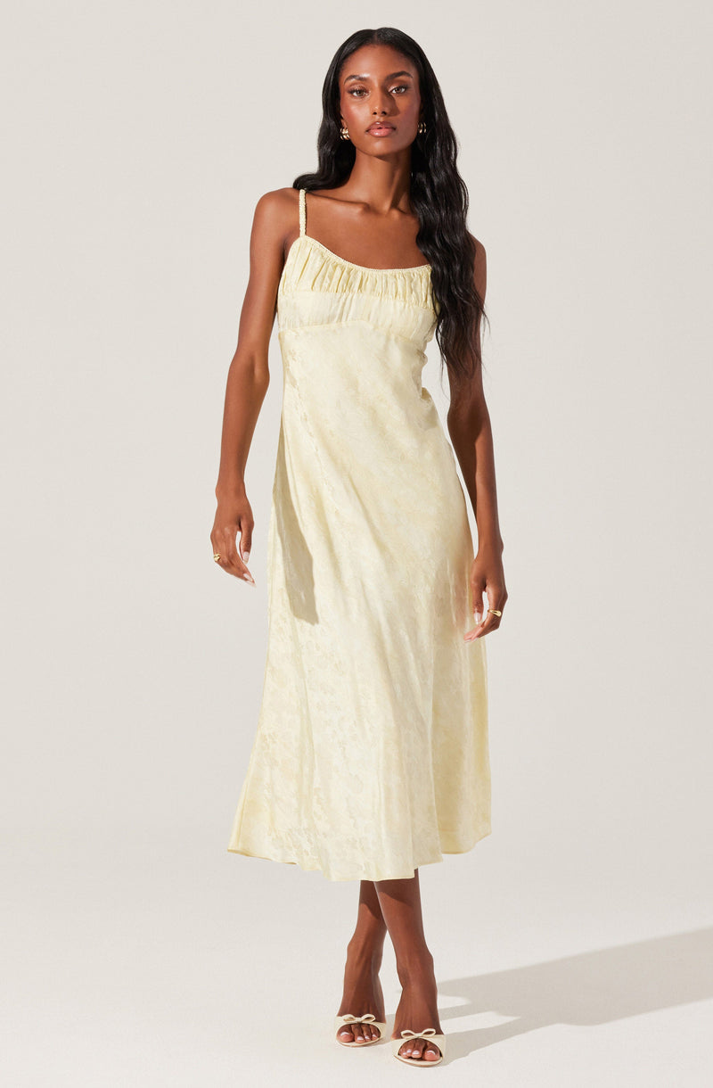 A woman is wearing a sleeveless, ankle-length dress with thin shoulder straps. The dress is made of a light, shimmery, pale yellow fabric with a subtle textured pattern. The top of the dress has a gathered detail across the bust. She is accessorized with gold hoop earrings and open-toed, heeled sandals.