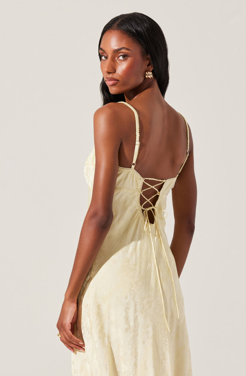 A woman is wearing a pale yellow, sleeveless dress with adjustable spaghetti straps. The back of the dress features a lace-up detail with a criss-cross pattern and a tied bow at the bottom. The fabric of the dress appears to have a subtle, textured pattern.