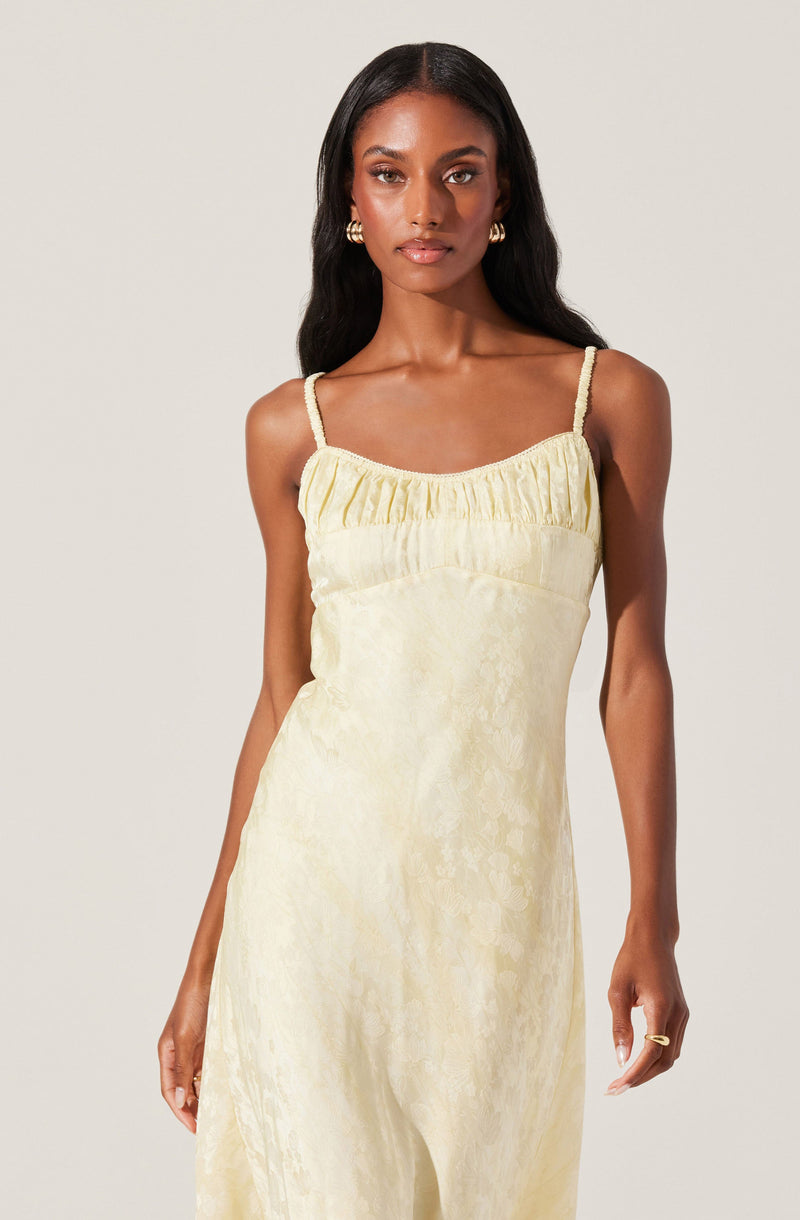 A woman is wearing a light yellow, silky slip dress with thin, braided spaghetti straps. The dress features a gathered detail along the neckline and has a subtle floral pattern or texture. She accessorizes with large, gold hoop earrings and a gold ring on her right hand.