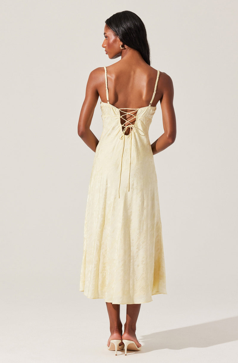 A woman is wearing a long, light yellow dress with a subtle floral pattern. The dress features spaghetti straps and a lace-up back with crisscrossing ties. The fabric appears to be lightweight and slightly textured. She is also wearing beige high-heeled shoes.