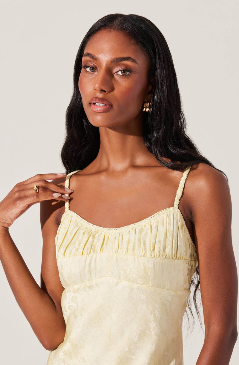 A woman is wearing a light yellow, sleeveless top with thin, ruched straps. The top has a gathered, pleated detail across the chest area. The fabric appears soft and has a subtle, textured pattern.
