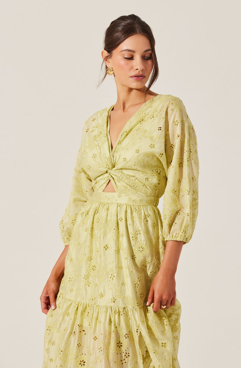 A woman is wearing a yellow eyelet lace dress with a V-neckline and long, puffed sleeves. The dress features a twist-front detail at the bust, creating a small cutout above the waist. The skirt is tiered with a ruffle at the hem, and the fabric has intricate floral patterns with eyelet cutouts throughout. She accessorizes with large, gold, geometric earrings.