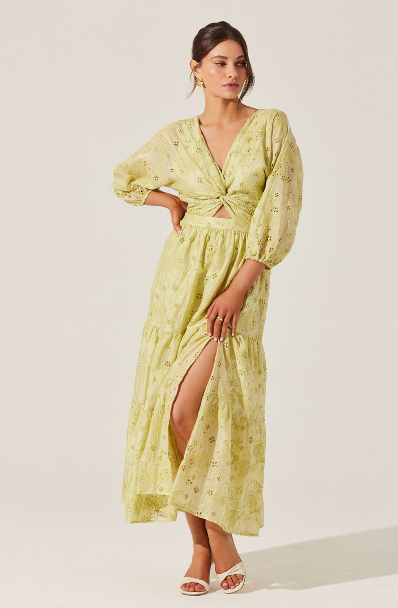 A woman is wearing a light yellow, ankle-length dress with eyelet embroidery. The dress has puffed, three-quarter length sleeves and a deep V-neckline with a twist knot at the front, creating a small cutout detail beneath the knot. The skirt features a high slit on one side, revealing her leg and a pair of white open-toe heels.