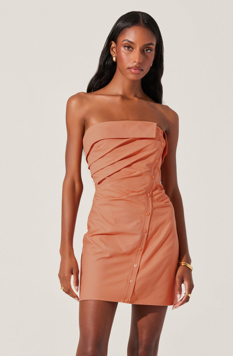 A woman is wearing a strapless, peach-colored mini dress with a asymmetrical, layered top section and button details running diagonally down the front. The fabric appears smooth and slightly shiny, giving a sleek and elegant look.