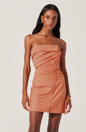 A woman is wearing a strapless, peach-colored mini dress with a asymmetrical, layered top section and button details running diagonally down the front. The fabric appears smooth and slightly shiny, giving a sleek and elegant look.