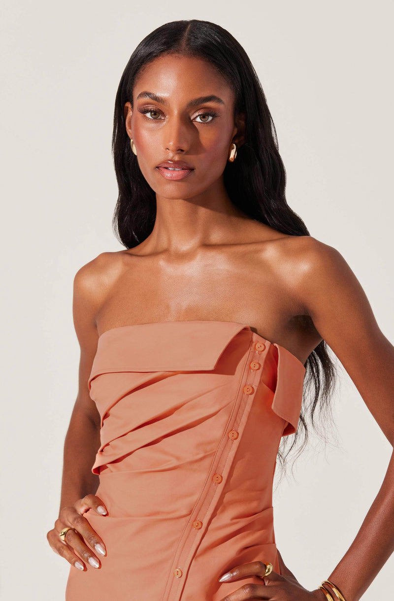 A woman is wearing a strapless peach-colored dress with a layered, pleated design on the bodice. The dress features a row of buttons running diagonally along the side. She has long, dark hair and is accessorized with gold hoop earrings, rings, and bracelets.