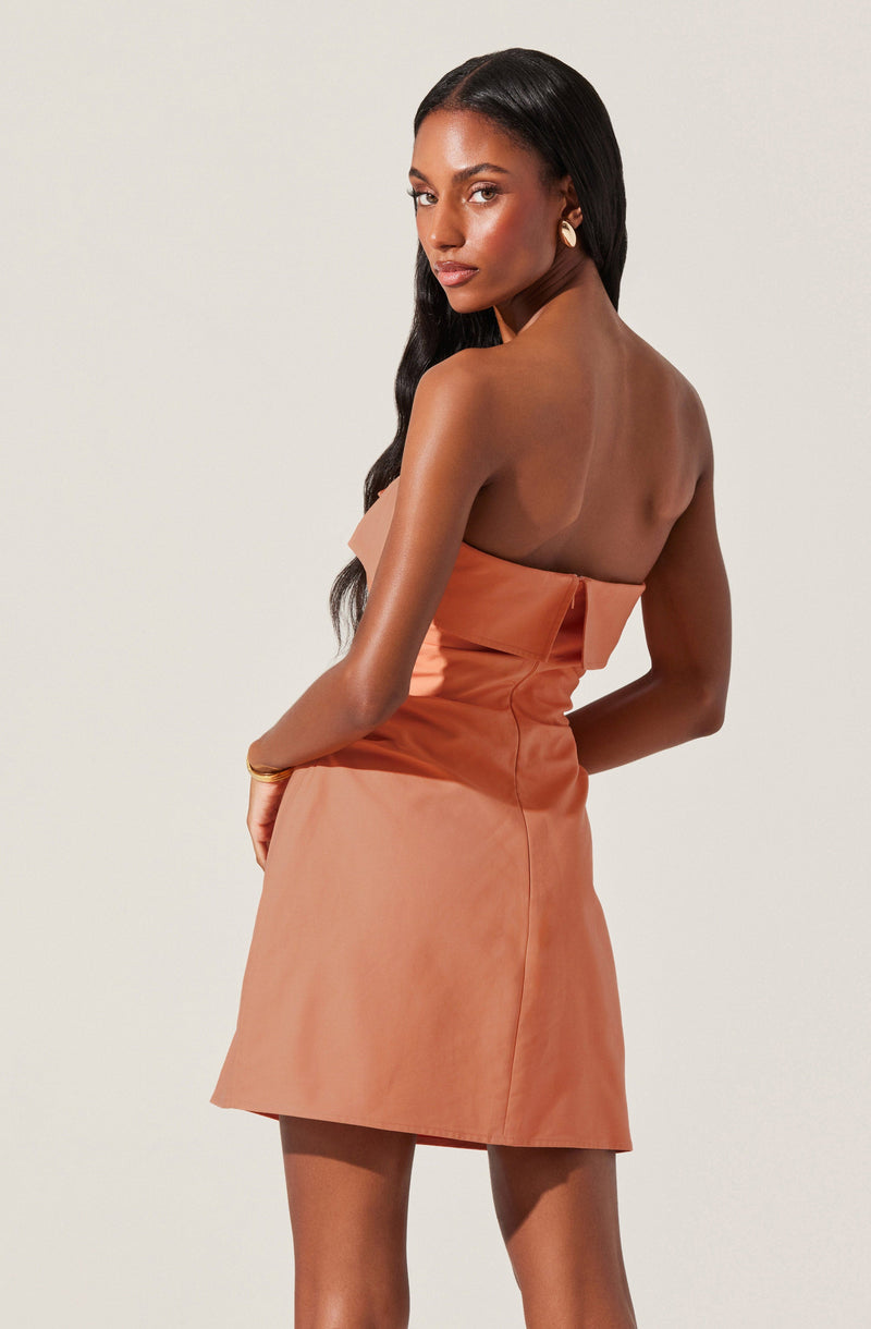 A woman is wearing a strapless, peach-colored dress that features a large bow detail on the front. The dress has a fitted bodice and a slightly flared skirt.