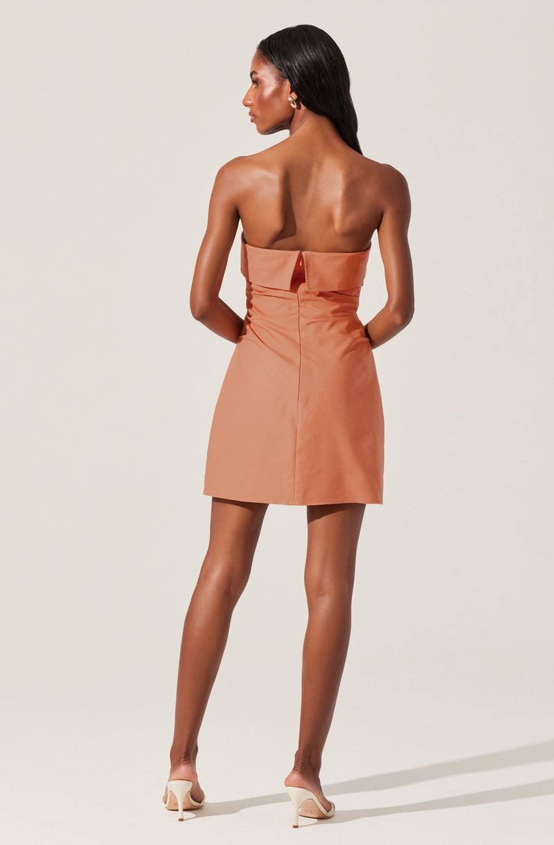 A woman is wearing a strapless, blush-colored mini dress with a wide waistband and a center back zipper. She has paired the dress with white open-toe heels.