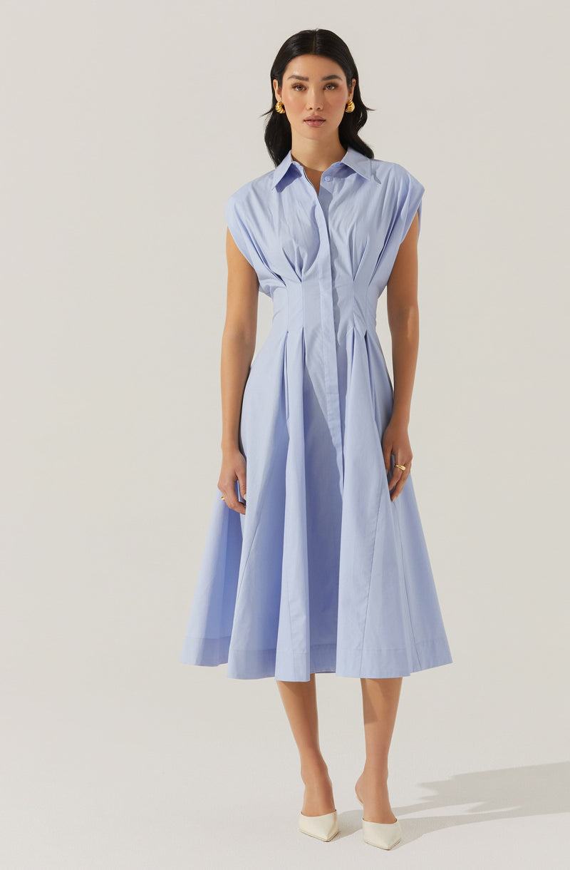 A woman is wearing a sleeveless, light blue, midi-length dress with a collared neckline and front pleats. The dress has a fitted waist and a full, flowing skirt. She is accessorized with gold earrings, a gold ring, and white pointed-toe heels.