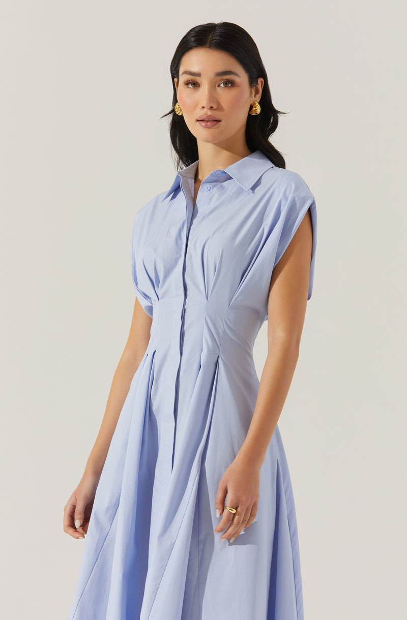 A woman wearing a light blue, sleeveless, knee-length dress with a collared neckline and a button-down front. The dress has pleats at the waist and side slits at the hem. She is accessorized with gold hoop earrings and a gold ring.