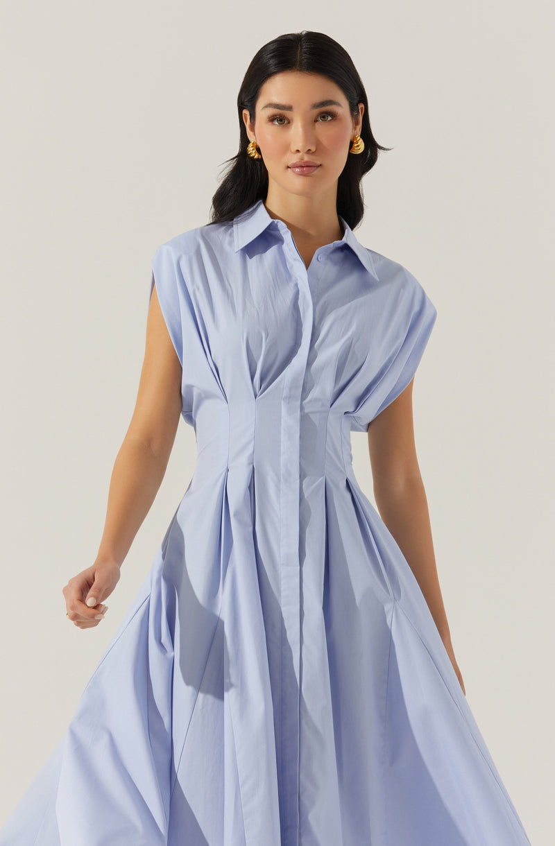 A woman is wearing a light blue sleeveless dress with a collared neckline and a button-down front. The dress features structured pleats and gathered fabric that creates a voluminous, flowy silhouette from the waist down.