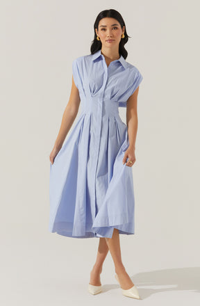 A woman is wearing a light blue shirt dress with short, capped sleeves and a classic collar. The dress features pleats running down the front, giving it a structured yet flowing appearance. She pairs the dress with off-white pointed-toe heels and gold earrings.