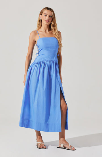 A woman is wearing a light blue sleeveless midi dress with thin spaghetti straps. The bodice is fitted, and the skirt is gathered with a side slit. The dress appears to have a lace-up detail on both sides of the bodice. She is also wearing white flat sandals.
