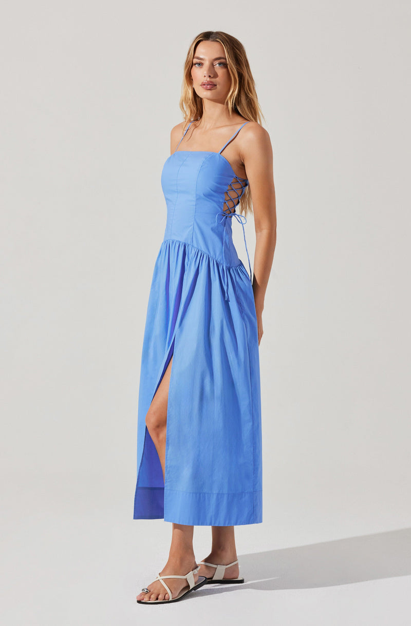 The dress is a sleeveless, light blue maxi dress with spaghetti straps. The bodice features a fitted, structured design with a corset-like appearance. On the sides, there are crisscrossing laces that tie up, creating a lattice effect. The skirt is gathered at the waist, flowing down to ankle length, and has a high slit on one side, exposing part of the leg. The fabric appears lightweight and slightly shiny. The dress is paired with white open-toe sandals.