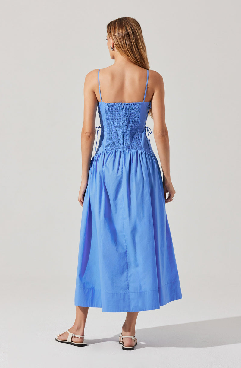 A woman is wearing a light blue dress with thin spaghetti straps. The top part of the dress is fitted with a ruched, smocked design, and the skirt is gathered and flows down to ankle length. The dress has ties on the sides at the waist. She is also wearing flat, open-toe sandals with ankle straps. Her hair is long and light brown.