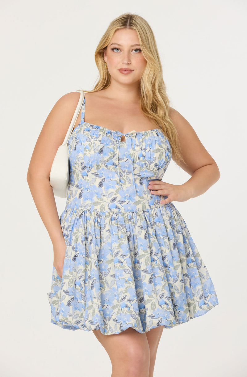 A woman wearing a sleeveless, light blue and cream floral dress with a fitted bodice featuring a small keyhole opening at the neckline and a gathered, full skirt. The dress has spaghetti straps and a subtle ruffle detail at the top.