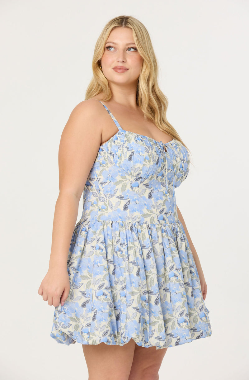 A woman is wearing a sleeveless, light blue dress with a floral pattern featuring shades of blue, beige, and green. The dress has thin spaghetti straps, a gathered neckline, and a ruffled, full skirt that falls above the knees.