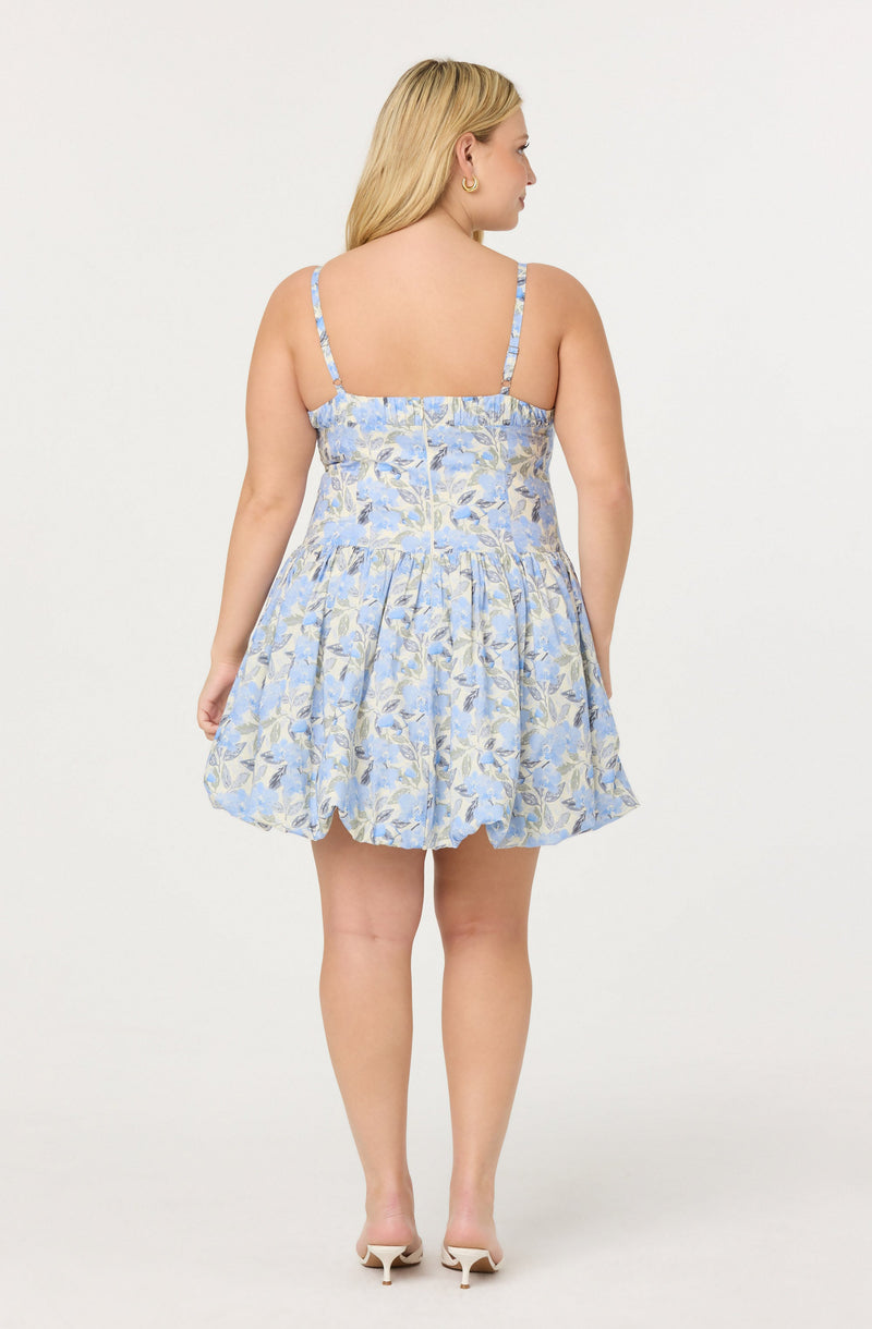 A woman is wearing a short, sleeveless dress with thin spaghetti straps. The dress features a light blue and beige floral pattern and has a fitted bodice with a slightly ruffled, gathered skirt. She is accessorized with small hoop earrings and white heels.