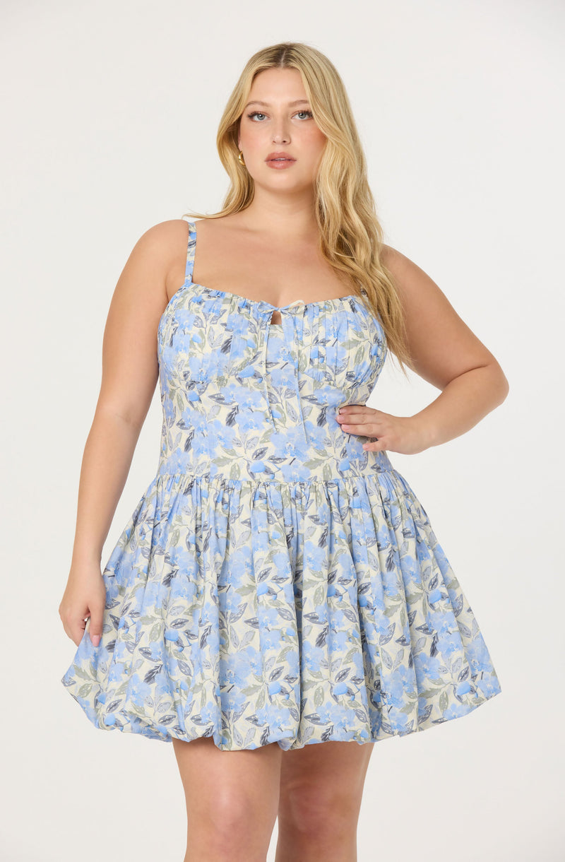 A woman is wearing a sleeveless dress with spaghetti straps. The dress has a fitted bodice with a small keyhole opening at the center and features a floral pattern in shades of blue, beige, and gray. The skirt is gathered and voluminous, creating a full, ruffled appearance.
