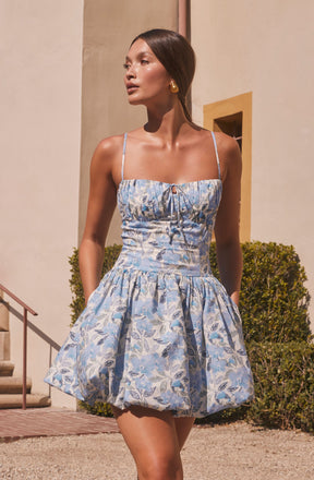 A woman is wearing a sleeveless dress with thin spaghetti straps. The dress has a fitted bodice with a small bow at the center and a flared, gathered skirt. The fabric features a light blue and white floral pattern with leaves and flowers.
