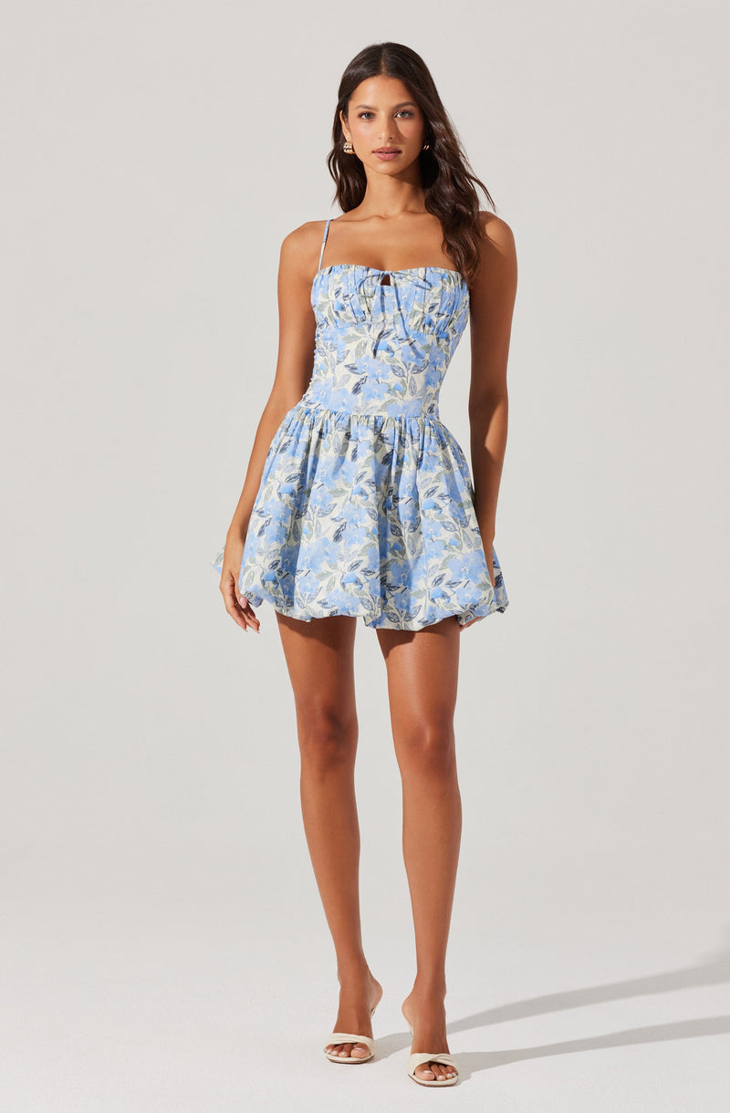 A woman is wearing a sleeveless, light blue dress with thin spaghetti straps and a fitted bodice with a keyhole cutout at the center. The dress features a floral pattern in shades of blue, white, and green, and has a gathered, flared skirt that falls mid-thigh. She is also wearing open-toed heels with ankle straps.