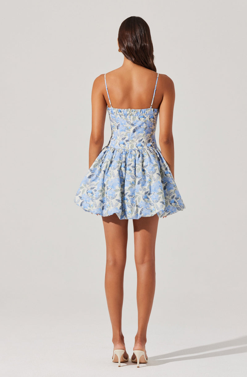 A woman is wearing a light blue dress with floral patterns. The dress has thin spaghetti straps and a fitted bodice that extends to a short, voluminous skirt. The fabric features a leafy design in shades of blue and gold. She is also wearing nude high-heeled shoes.