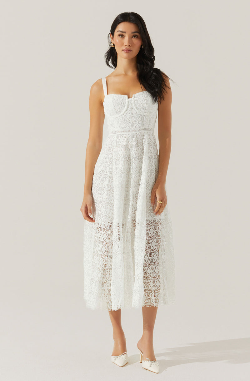 The woman is wearing a white, sleeveless, lace dress with a fitted bodice and straps over her shoulders. The dress features a sheer, patterned lace fabric that extends to mid-calf length, with a lining underneath only in the bodice area, leaving the lower part semi-transparent. She is also wearing white heeled shoes with pointed toes and decorative bows.
