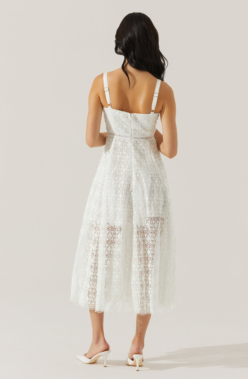 A woman is wearing a white, sleeveless dress with adjustable straps. The dress features an intricate lace pattern with geometric and floral motifs, and it extends to mid-calf length. She is also wearing white high-heeled shoes.